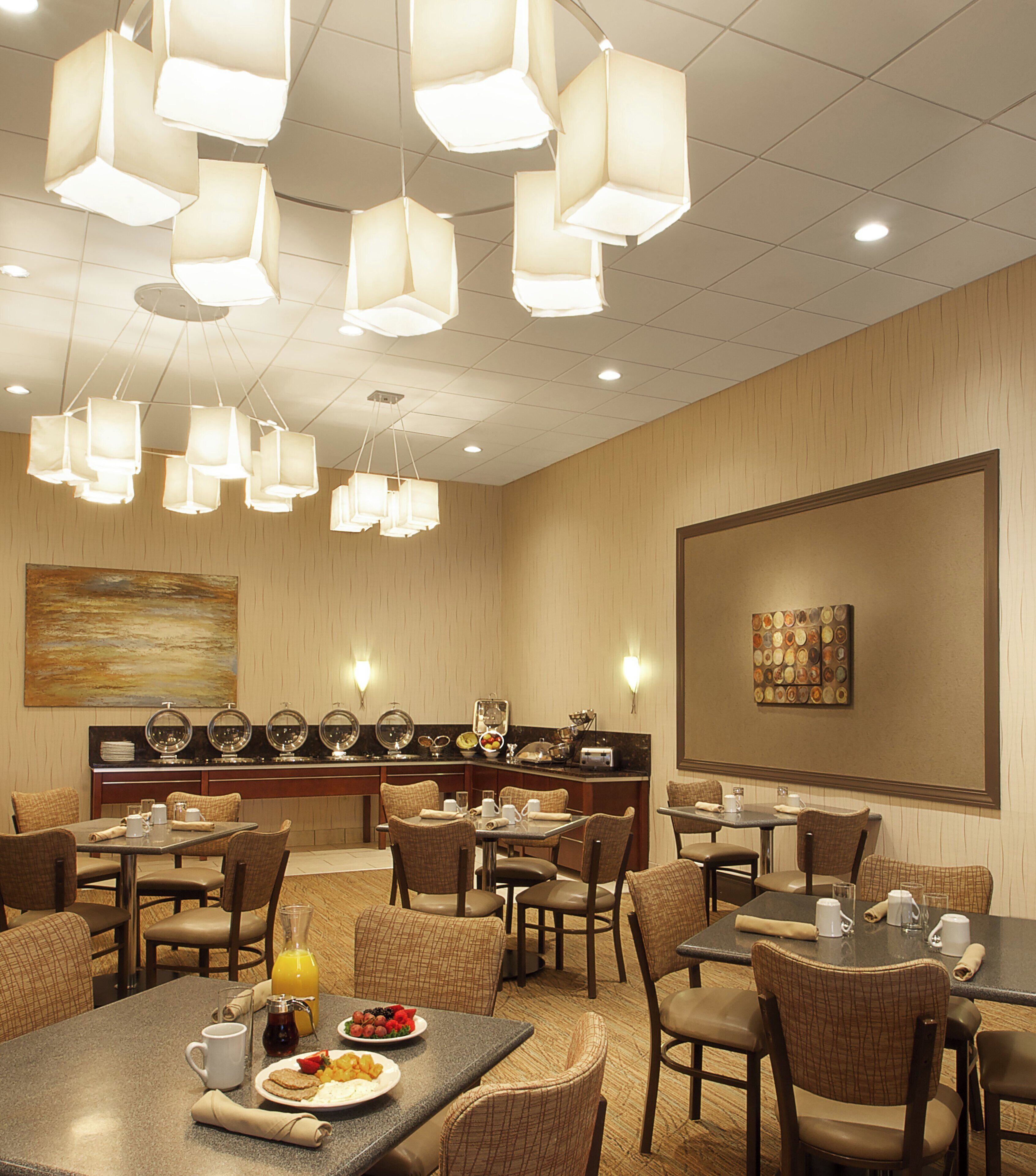 Un'area ristorazione di un hotel con diversi tavoli e sedie, illuminazione a soffitto e un grande quadro appeso al muro.