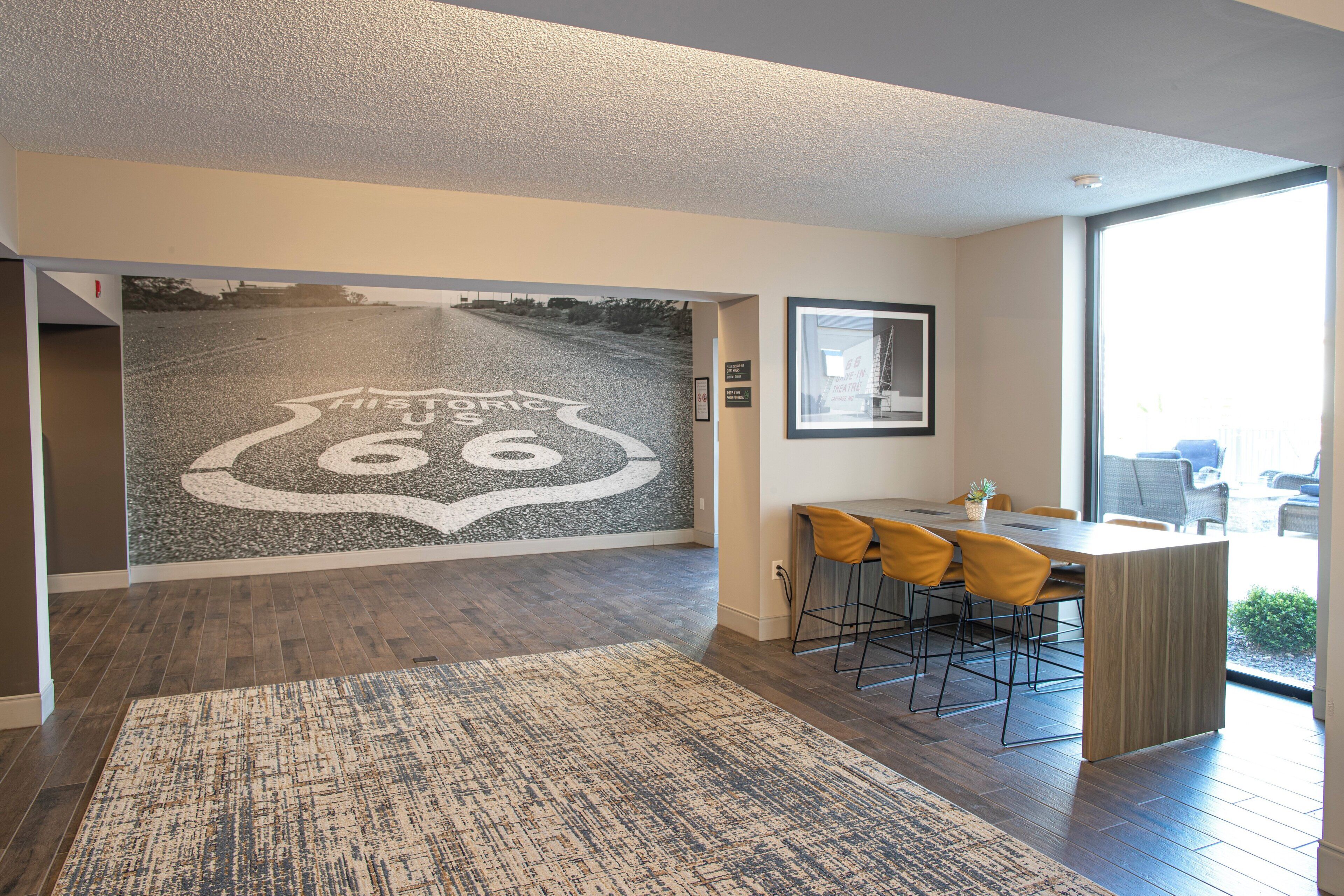 Un intérieur moderne agrémenté d’une grande fresque murale représentant la Route 66, d’un coin repas en bois avec des chaises jaunes et d’une grande fenêtre offrant une vue sur l’extérieur.