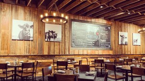 Ein Restaurant Innenraum mit holzverkleideten Wänden, hängenden Leuchten und einer Tafel mit der Speisekarte. Tische und Stühle sind zum Essen angeordnet.