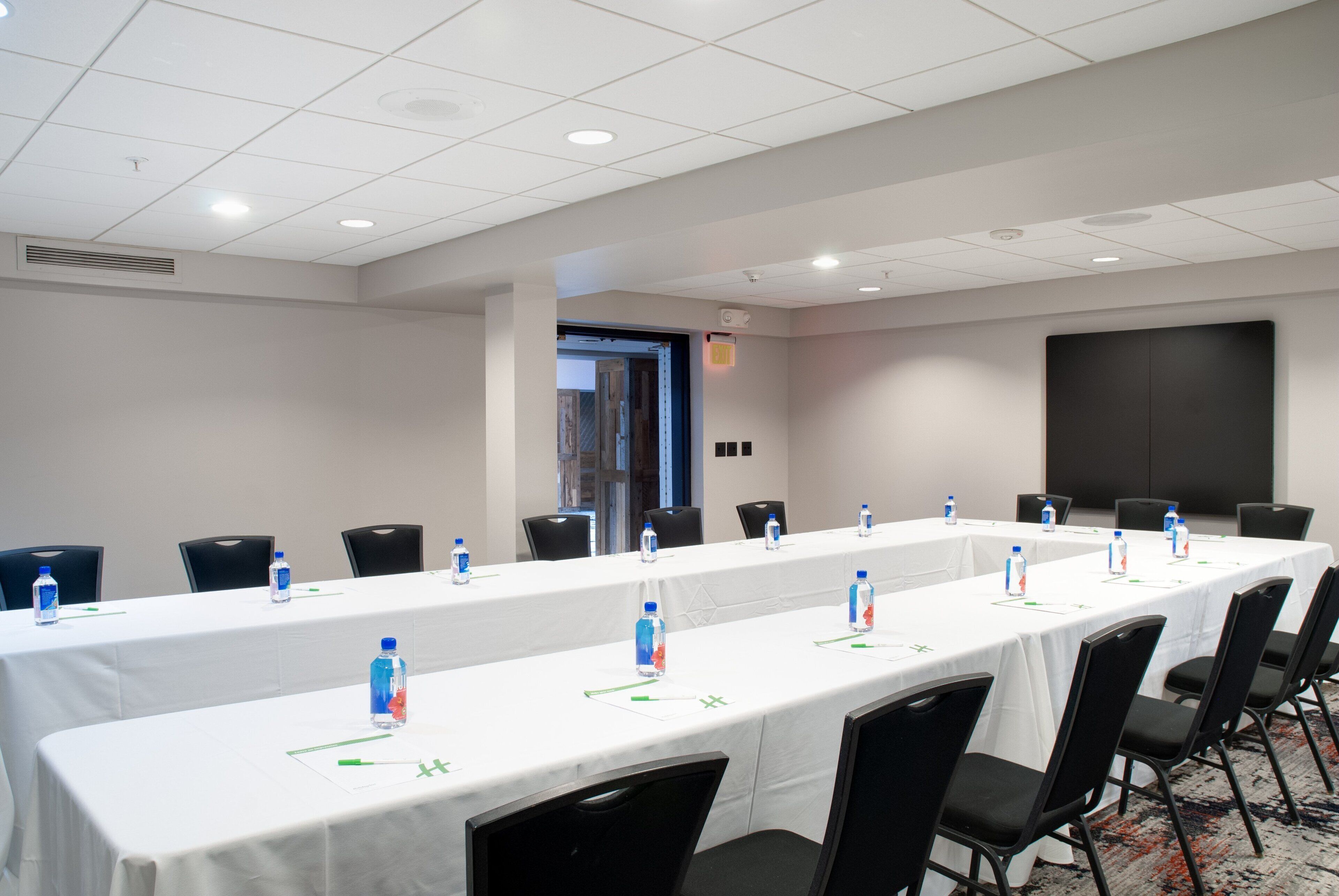 Une salle de conférence avec une longue table, des chaises noires, des bouteilles d’eau et un écran de projection.