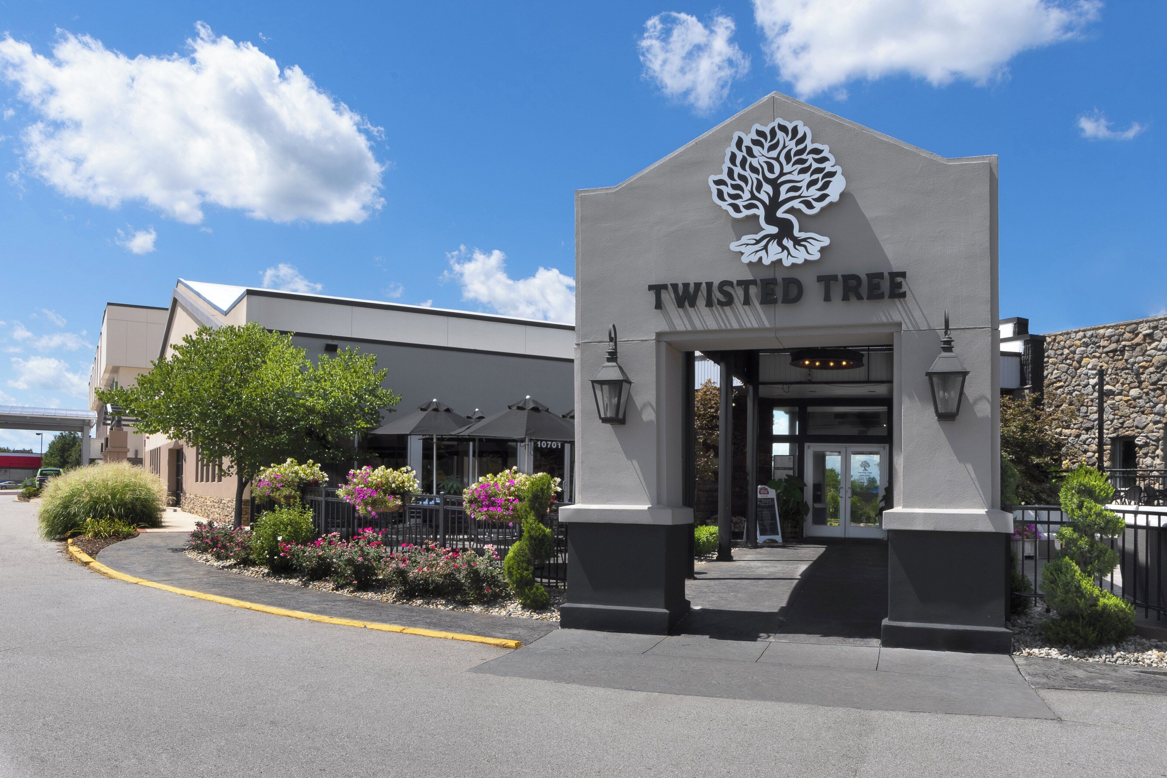 Un edificio con un letrero que dice 'TWISTED TREE' y un logotipo de árbol. Hay mesas y sillas al aire libre, y un área de acceso pavimentada.