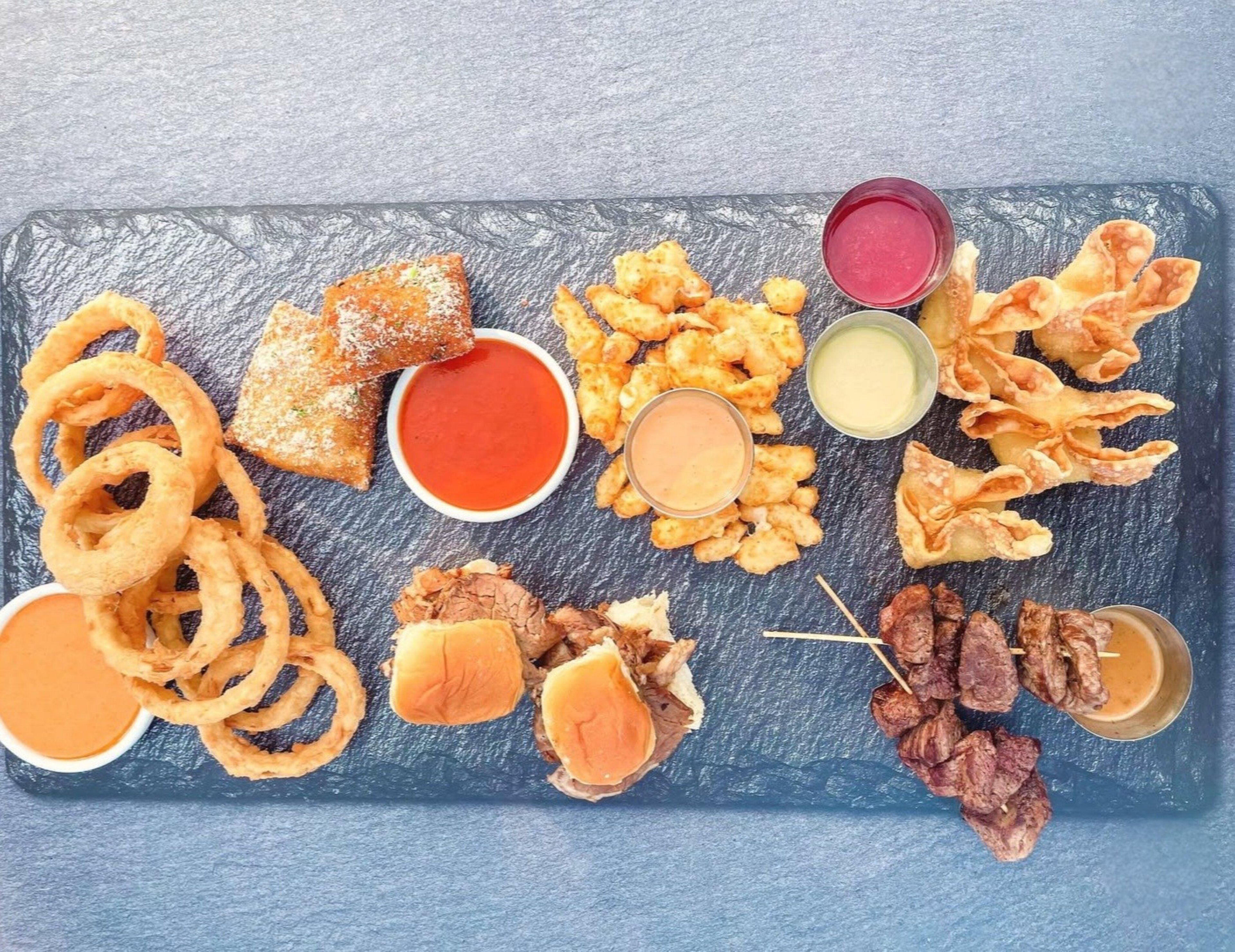 Variedad de alimentos fritos como aros de cebolla, hamburguesas y brochetas de carne, servidos sobre una tabla de pizarra con diferentes salsas para mojar.