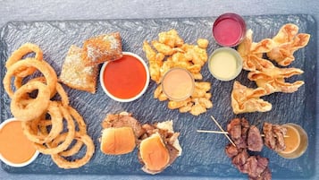 Variedad de alimentos fritos como aros de cebolla, hamburguesas y brochetas de carne, servidos sobre una tabla de pizarra con diferentes salsas para mojar.