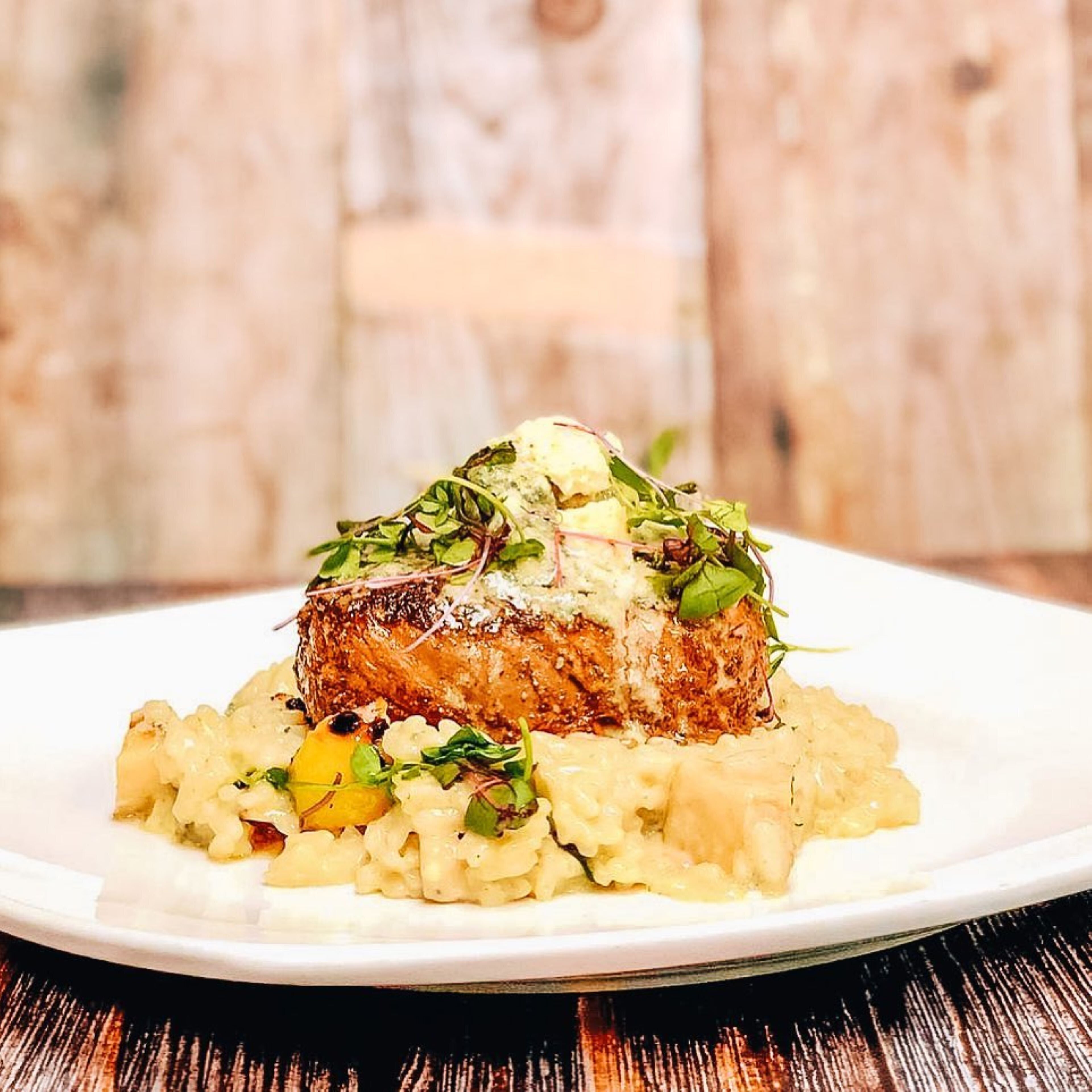 Un plato con un trozo de carne, posiblemente cerdo, cubierto con salsa cremosa y adornado con vegetales, servido sobre puré de papas.