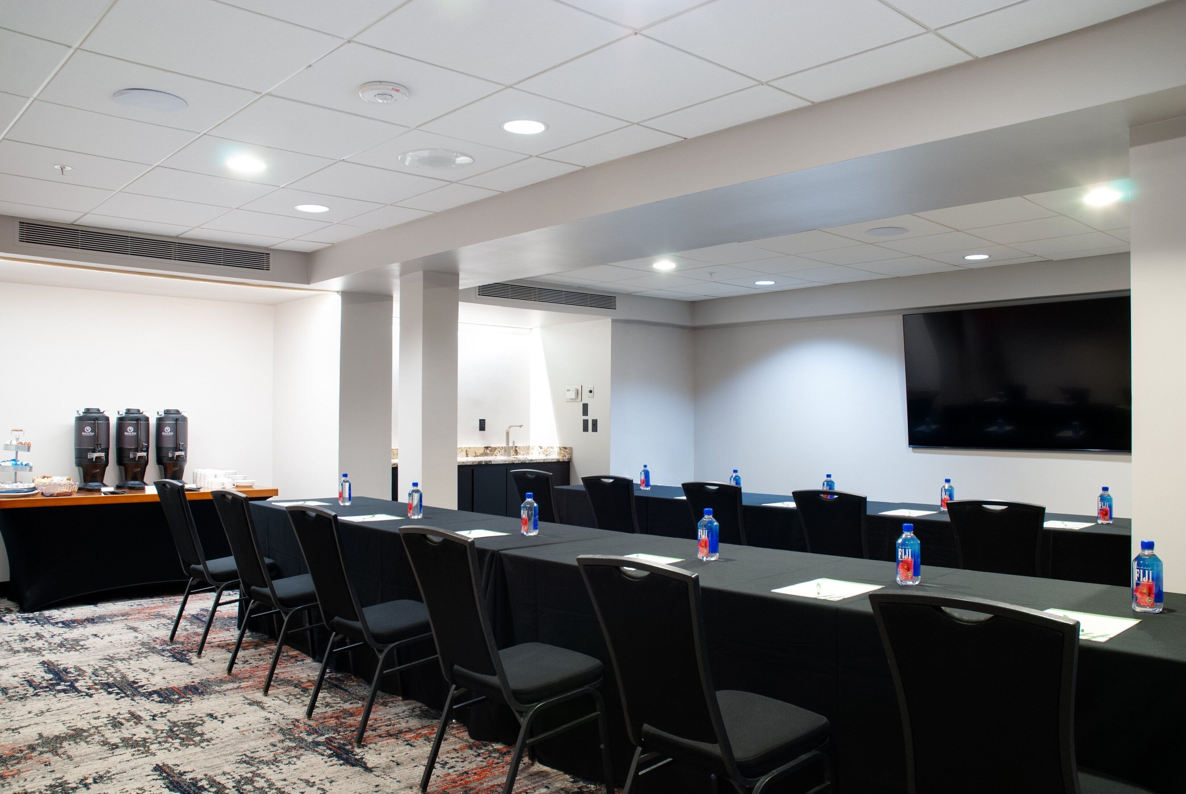 Un salón de conferencias con una larga mesa, sillas, botellas de agua, una pantalla de proyector y un rincón de café.