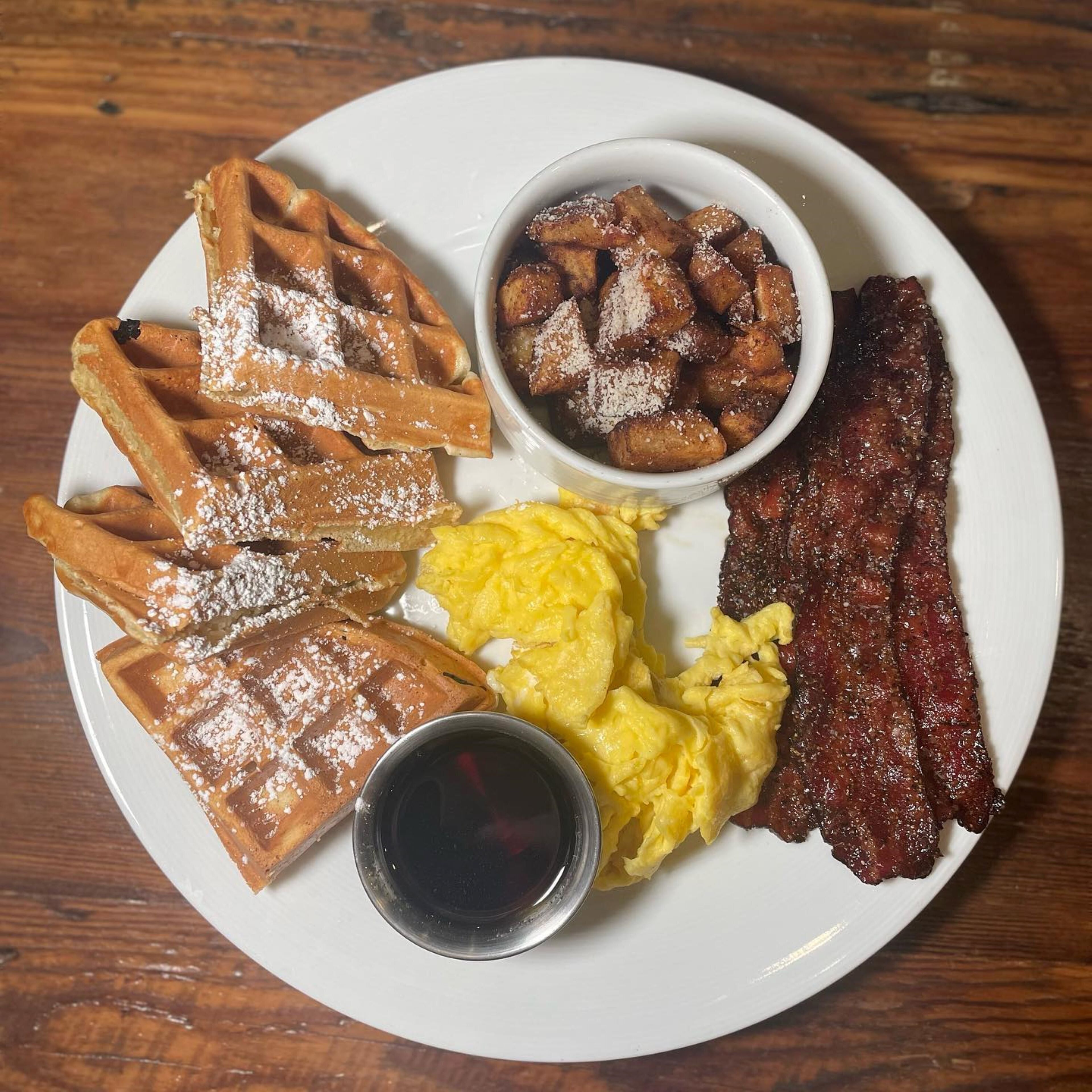 A plate with waffles, scrambled eggs, bacon, and a small bowl of what appears to be French toast.