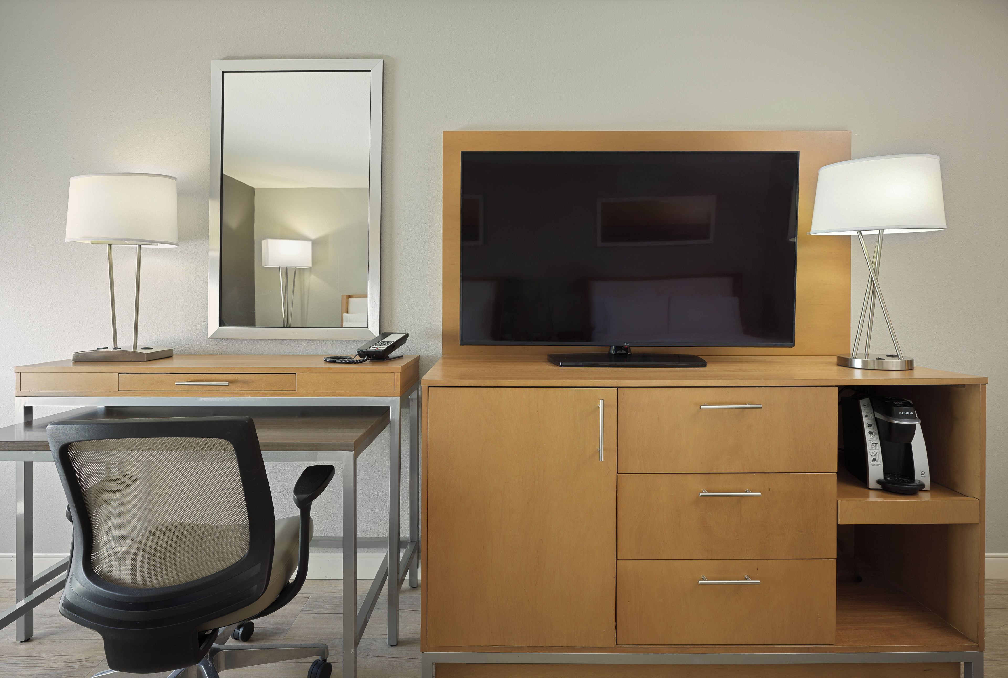 Habitación de hotel con un escritorio de madera, televisión, dos lámparas de noche, una silla y una cafetera.