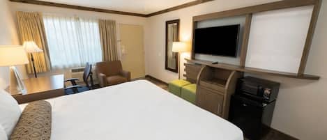 Una habitación de hotel con una cama grande, un escritorio con lámpara, una silla, un televisor montado en la pared y un refrigerador pequeño.