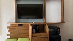 A room with a flat-screen TV mounted on a wooden wall unit, a coffee machine, a microwave, and a green chair. - The Dalles Inn (The Dalles)