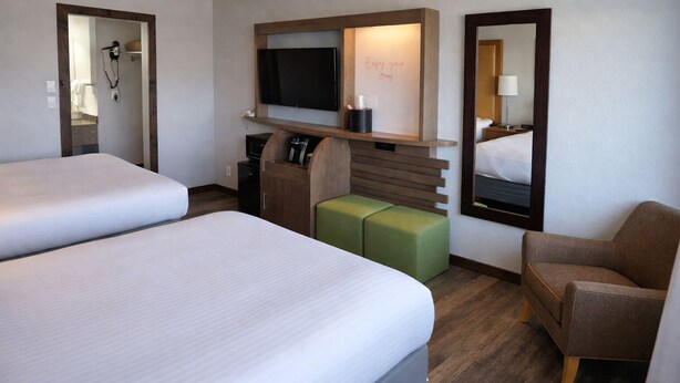 A hotel room with two beds, a TV, a desk with a computer, a mirror, and a green ottoman.