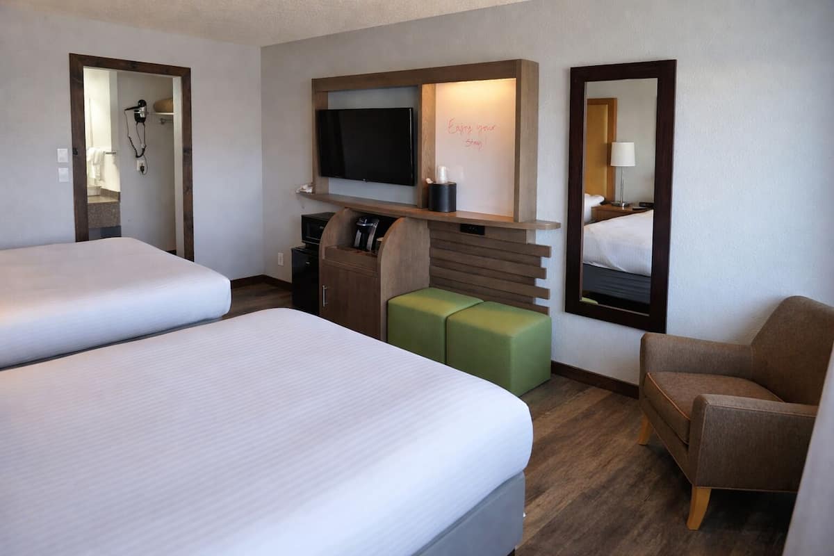Habitación de hotel con dos camas, un televisor, un escritorio con computadora, un espejo y un reposapiés verde.