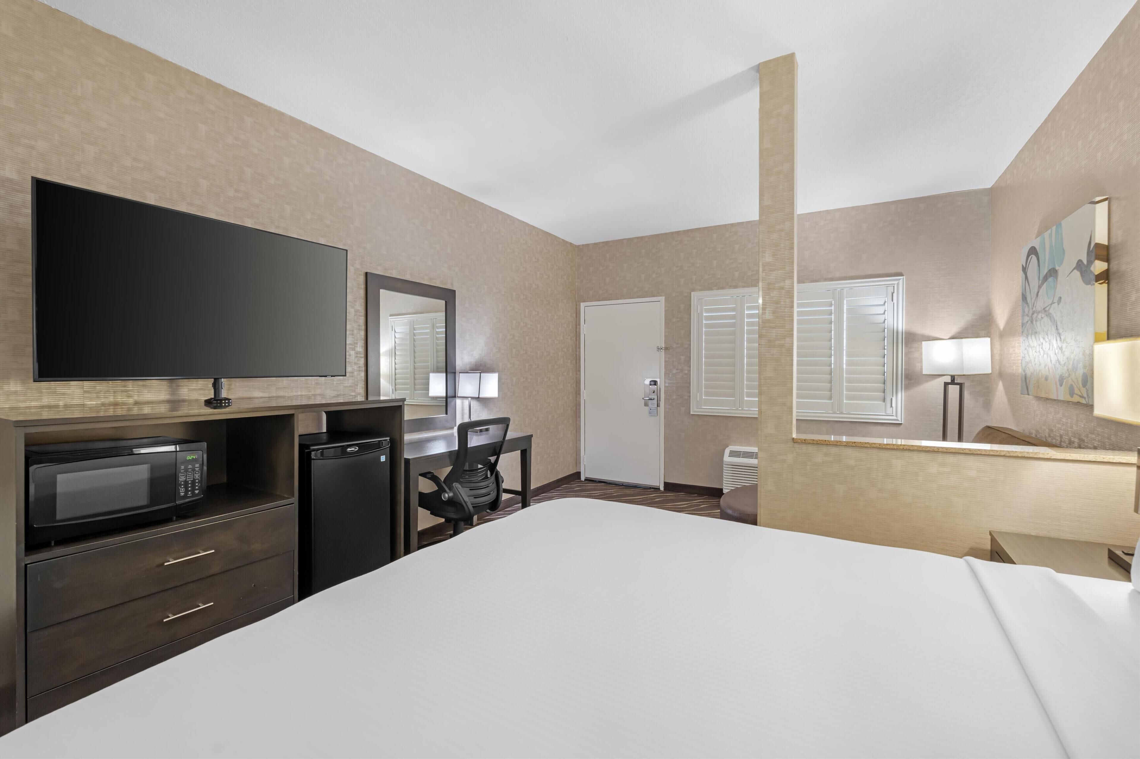 A hotel room with a large bed, a flat-screen TV, a microwave, a refrigerator, a desk with a chair, and a door with a window.