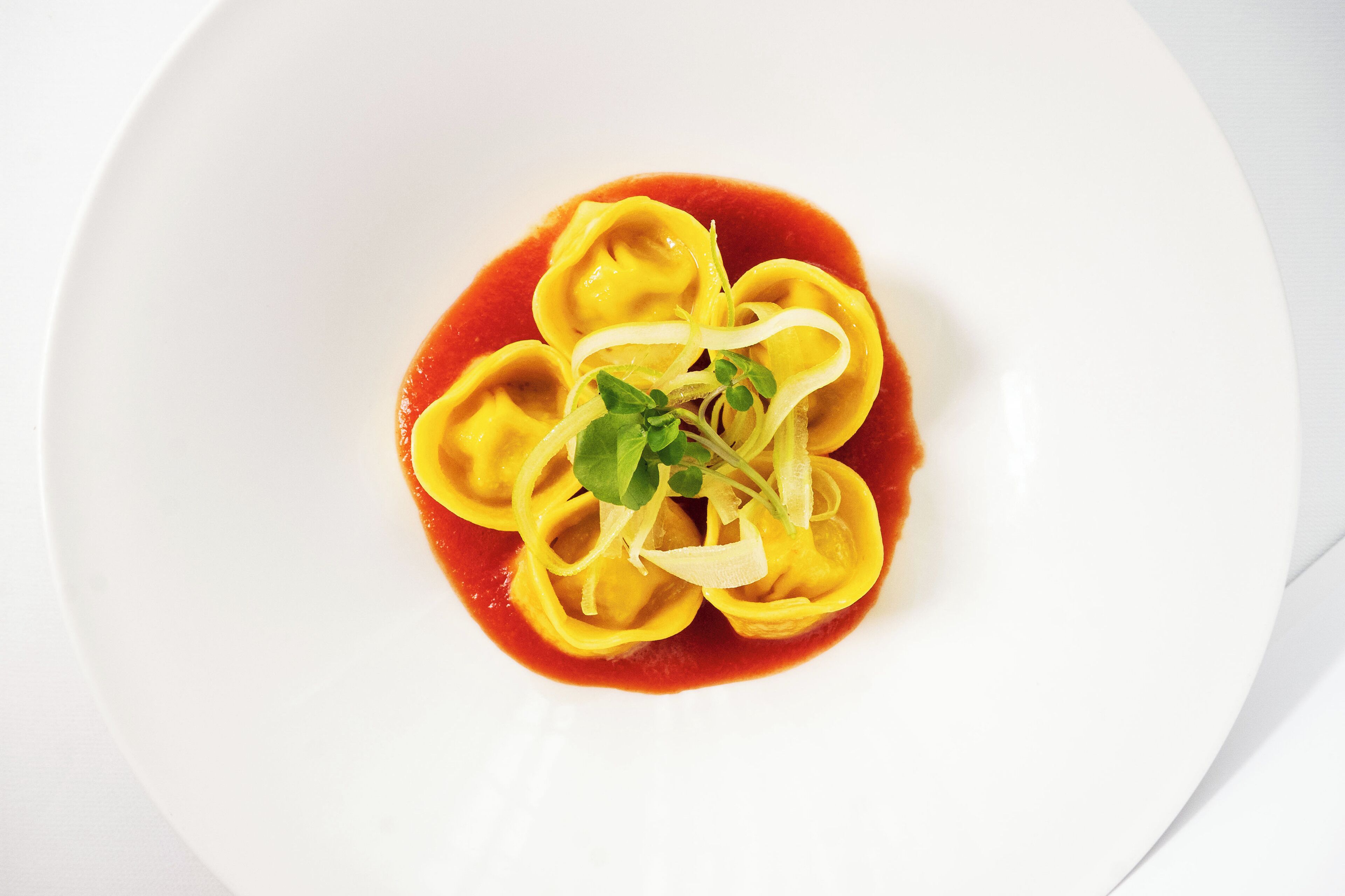 Un plato blanco con ravioles en salsa roja, adornado con brotes tiernos y queso rallado.