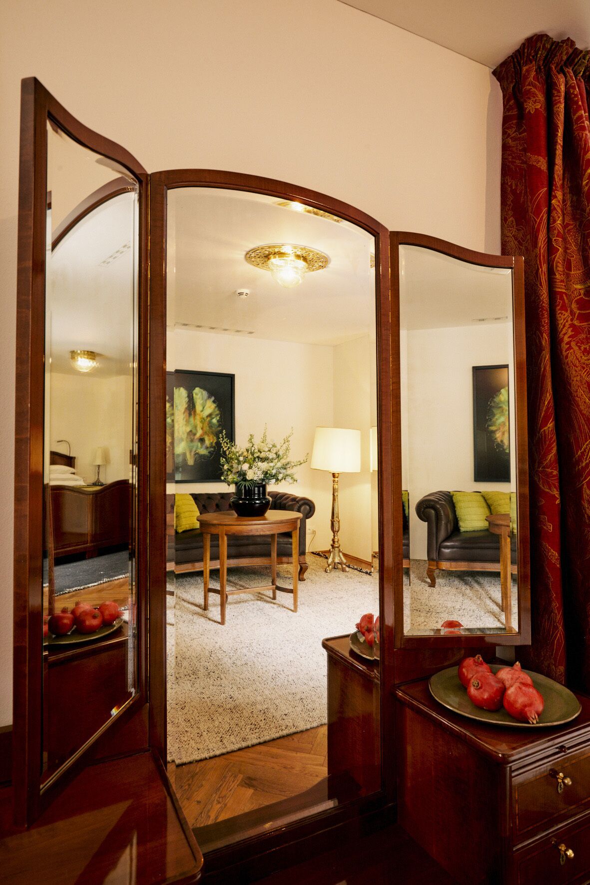A room with a wooden floor, a sofa, a table with a vase, a lamp, and a mirror reflecting the room.