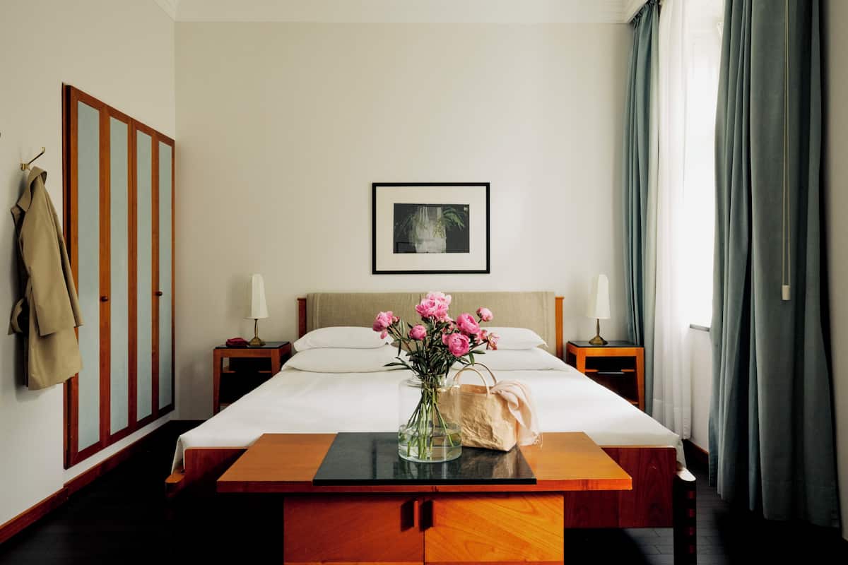 Ein Hotelzimmer mit Bett, Nachttisch mit Blumen in einer Vase, Lampe, Kleiderhaken und einem gerahmten Bild an der Wand.