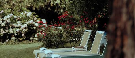 A row of white lounge chairs with towels, set on a grassy lawn, surrounded by dense foliage and flowering plants.