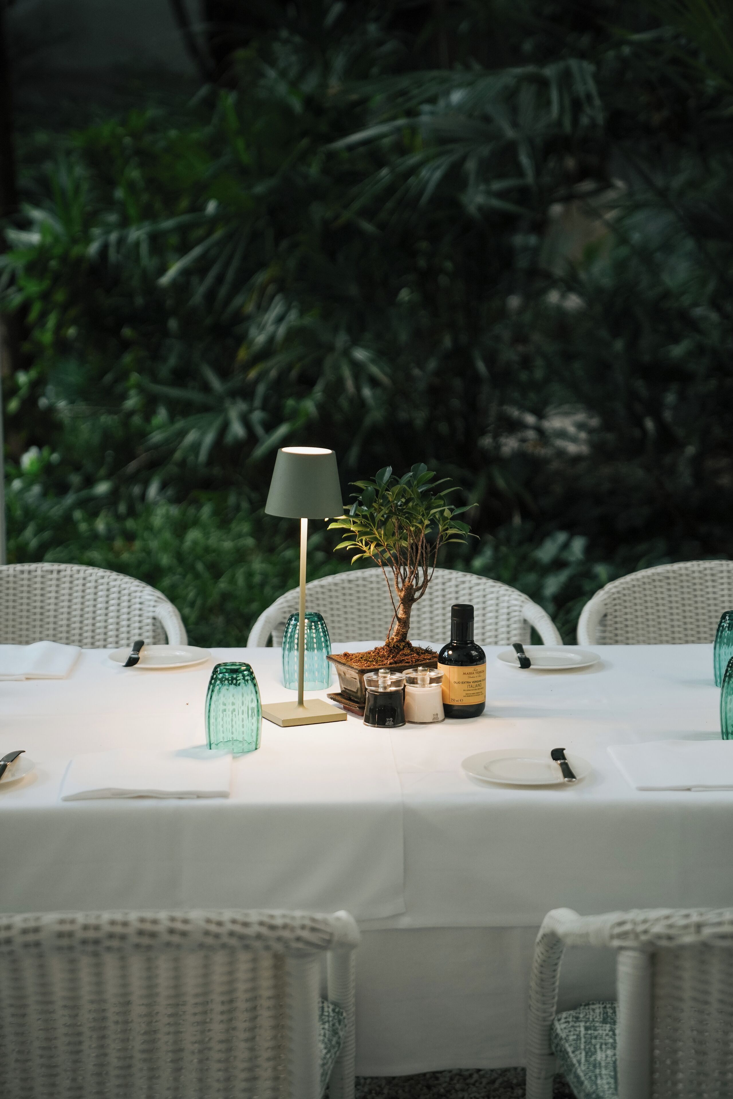Una mesa lista para comer, con una lámpara, una planta pequeña y varios objetos sobre un mantel blanco.