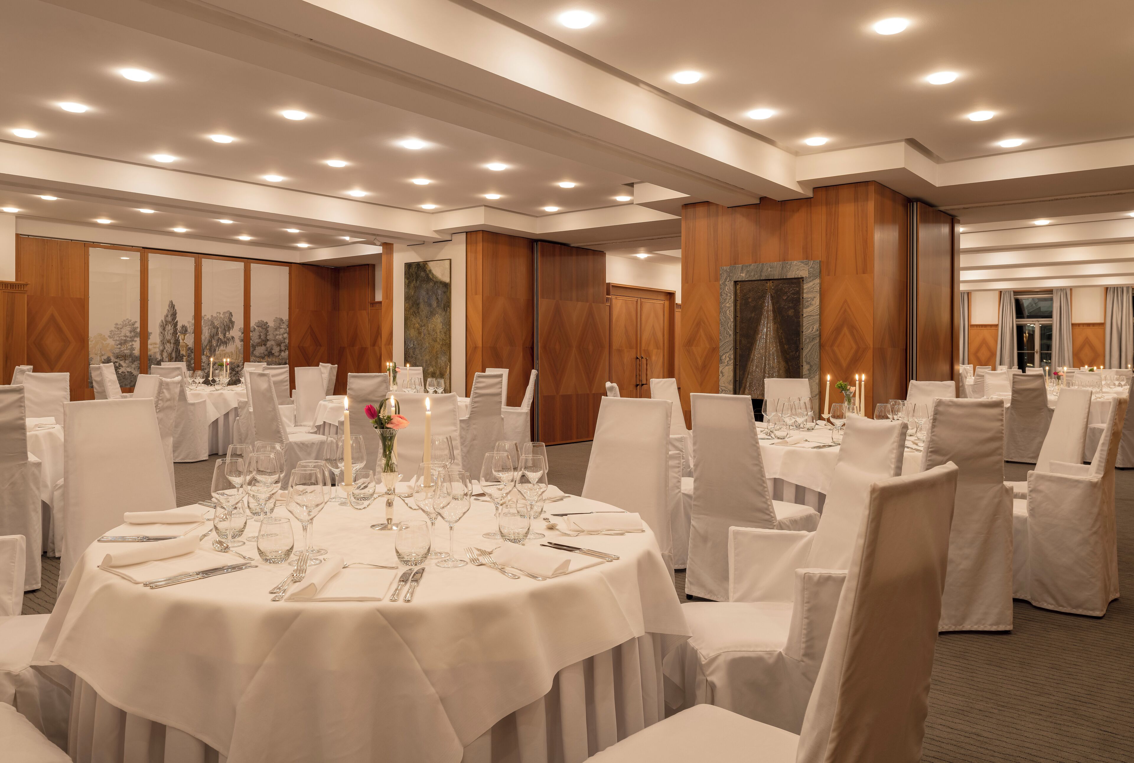 A banquet hall with round tables set for dining, white chairs, and a polished wooden finish.
