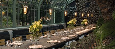 A long dining table set with chairs, candles, and floral centerpieces in a greenhouse with a stone wall and glass ceiling.