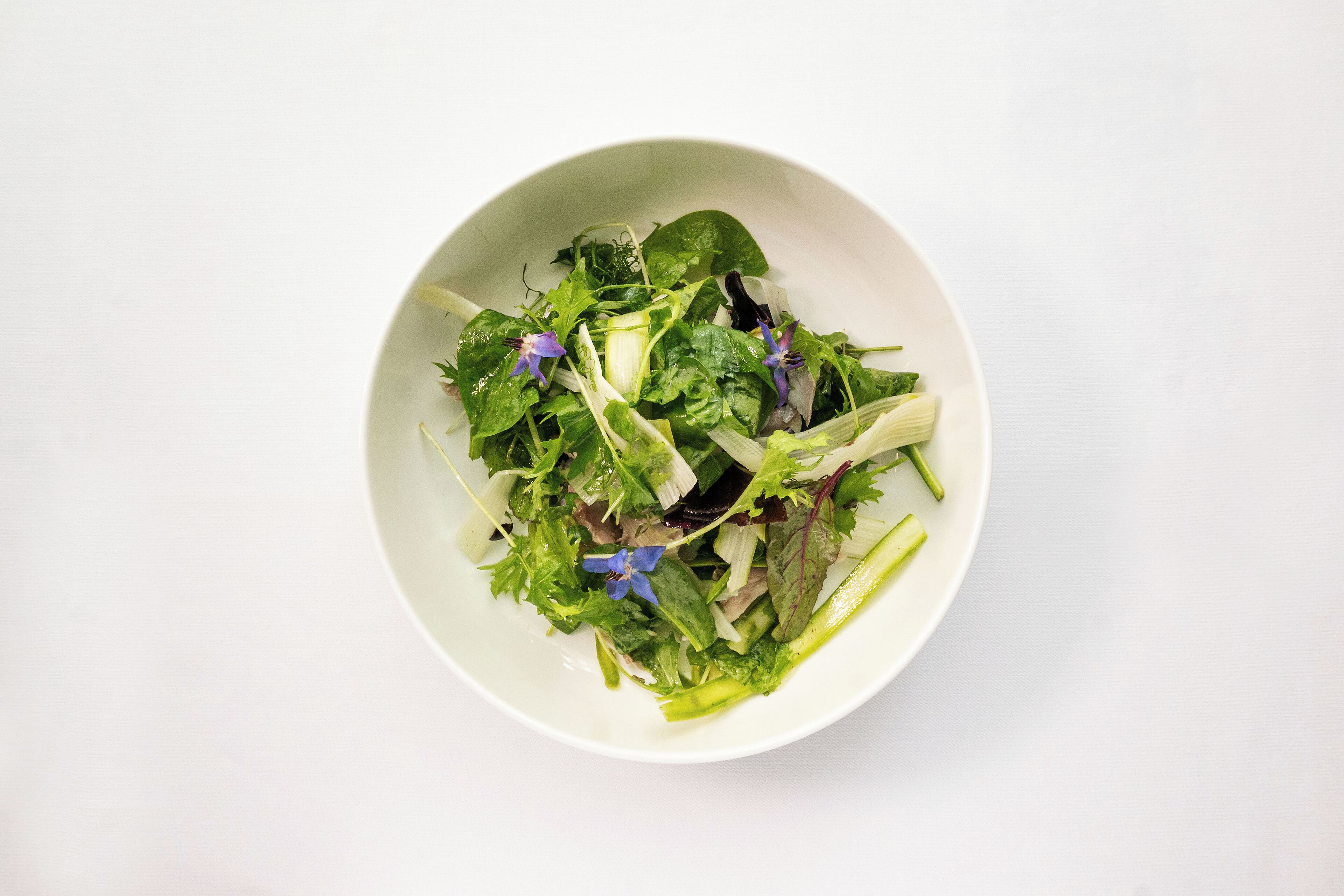 Un tazón blanco con una ensalada de diferentes vegetales de hoja y flores moradas.
