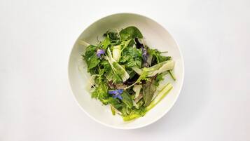Un tazón blanco con una ensalada de diferentes vegetales de hoja y flores moradas.