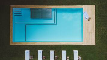 Una piscina rectangular con una terraza de madera y una zona sombreada con sombrillas.