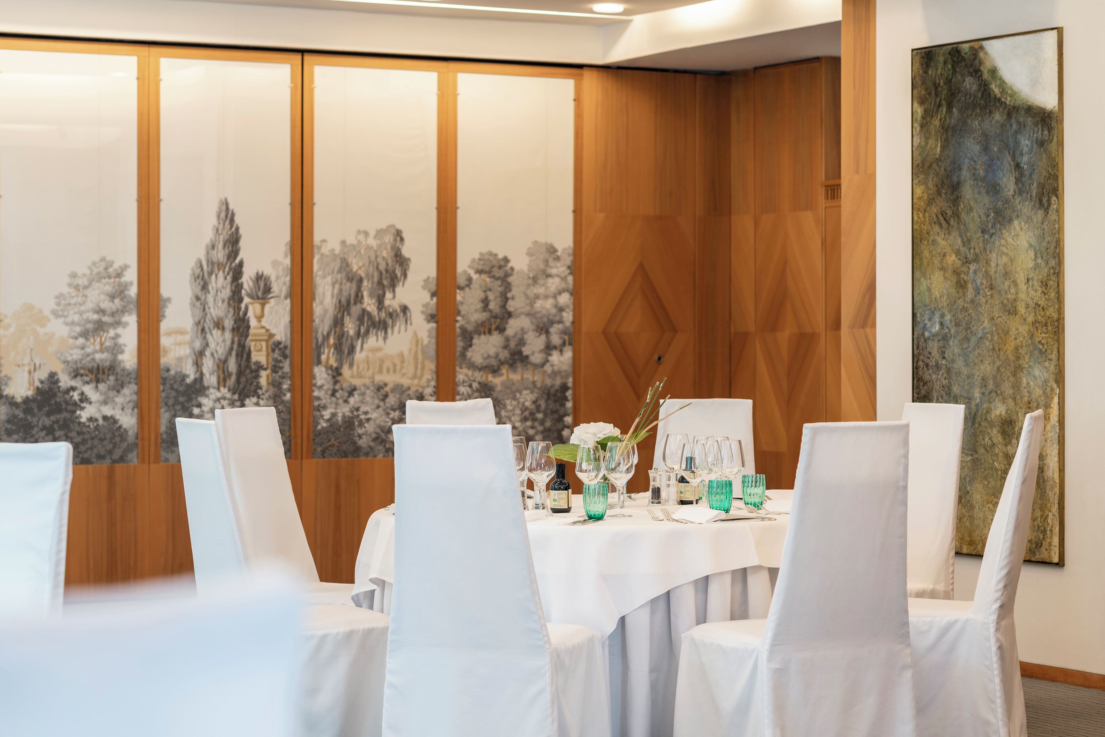 A dining area with a round table set for six, white chairs, and a backdrop of a traditional mural.