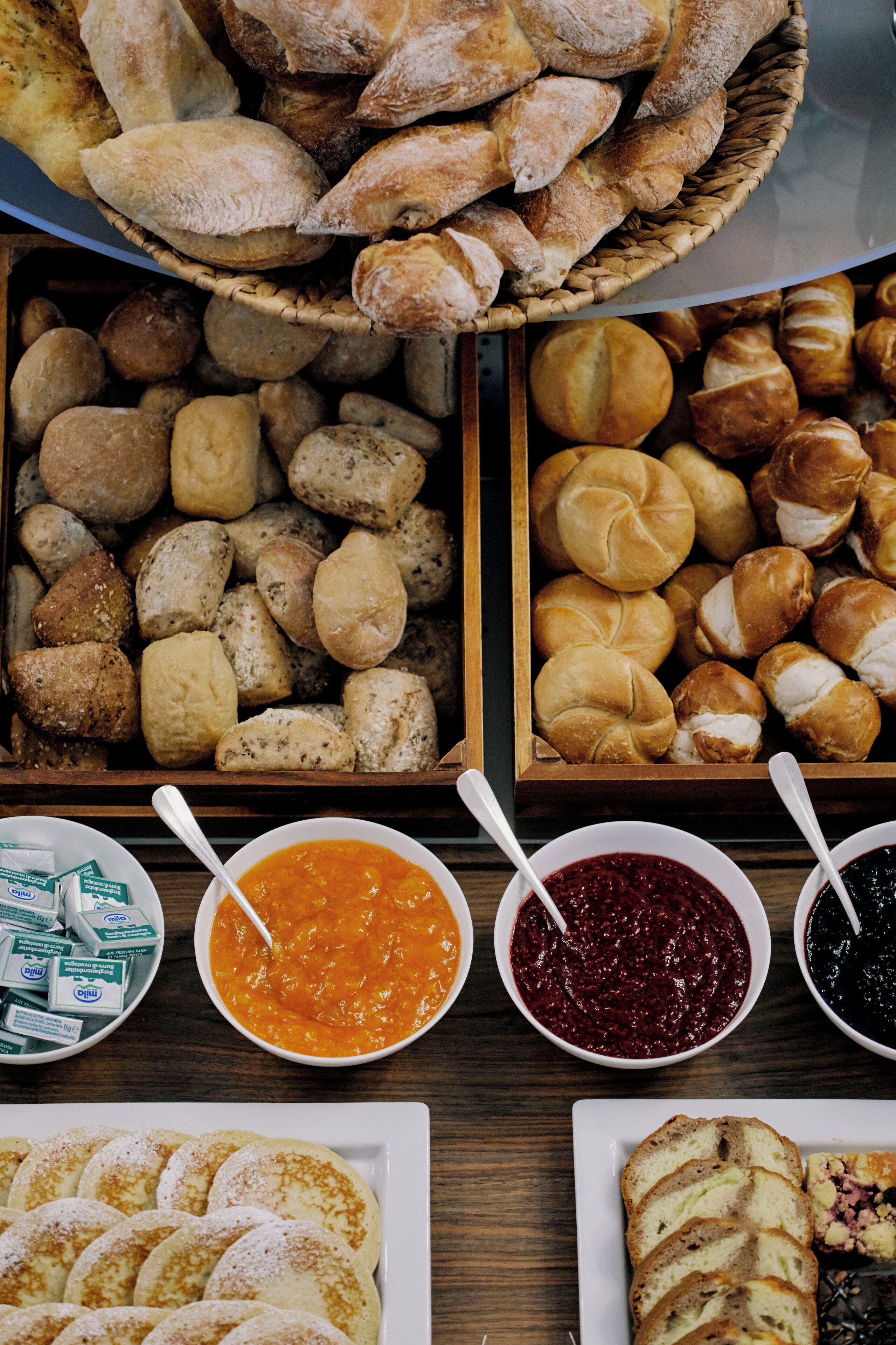 Una variedad de productos de panadería, como pan, bollos y pasteles, junto con recipientes de mermelada y mantequilla.