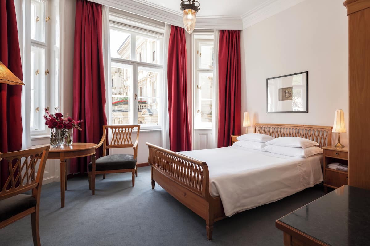 Ein Hotelzimmer mit einem großen Bett, einem Holzkopfstück, zwei Stühlen, einem kleinen Tisch mit Blumenvase und einem Fenster mit roten Vorhängen.