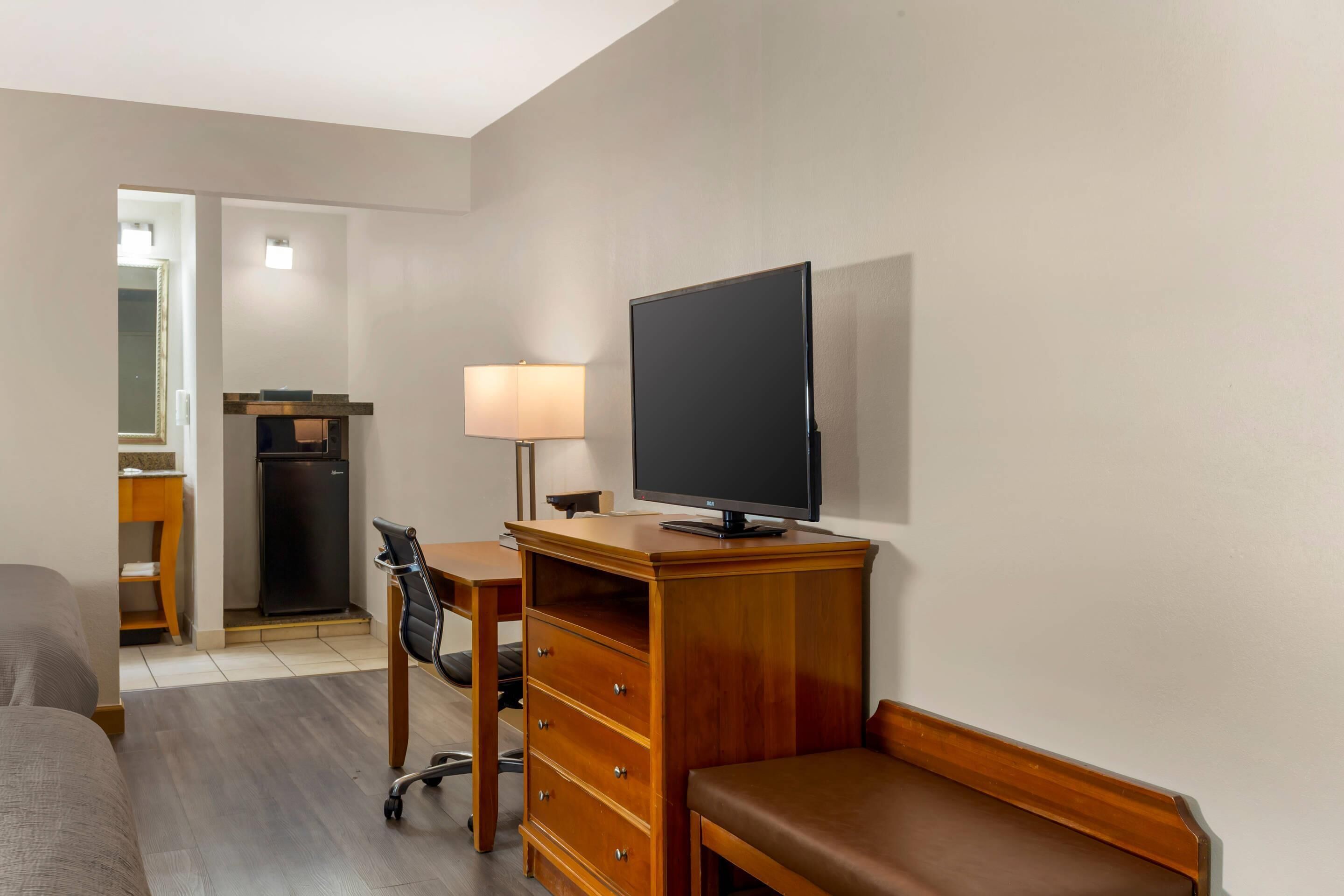 A hotel room with a bed, a desk with a television, a chair, a lamp, and a refrigerator.