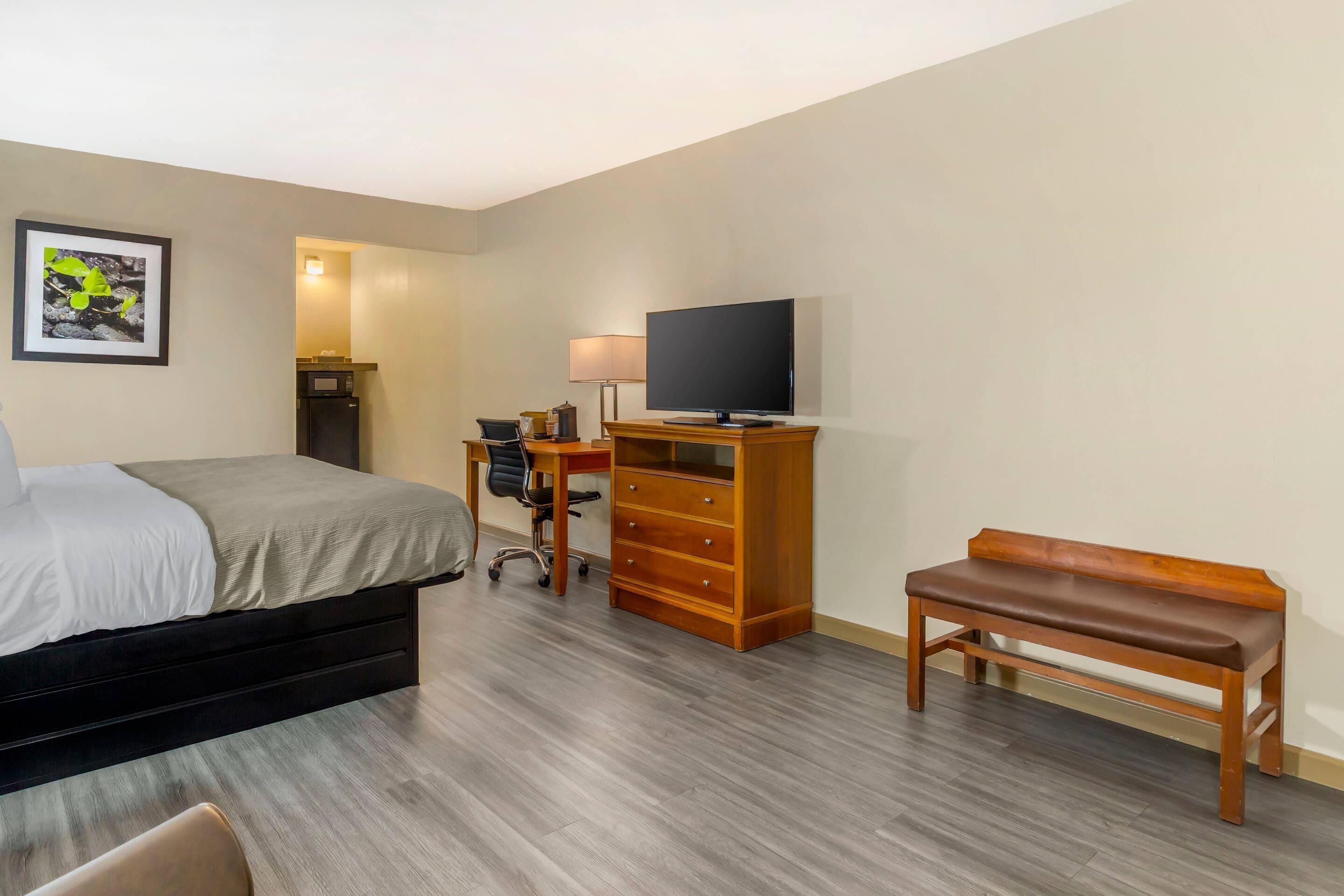 Habitación de hotel con cama, escritorio con ordenador, televisor, banco de madera y cómoda.