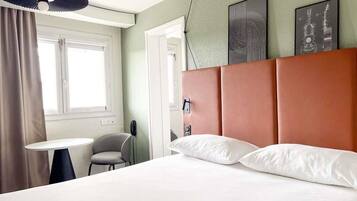 Una habitación de hotel con una cama, una silla, una mesita, una ventana con cortinas y una obra de arte en la pared.