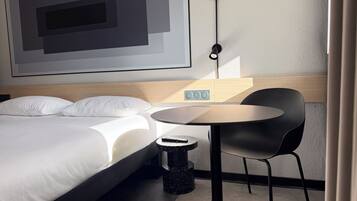 Ein modernes Hotelzimmer mit einem Bett, einem runden Tisch, einem schwarzen Stuhl und einer an der Wand montierten Steuerung.