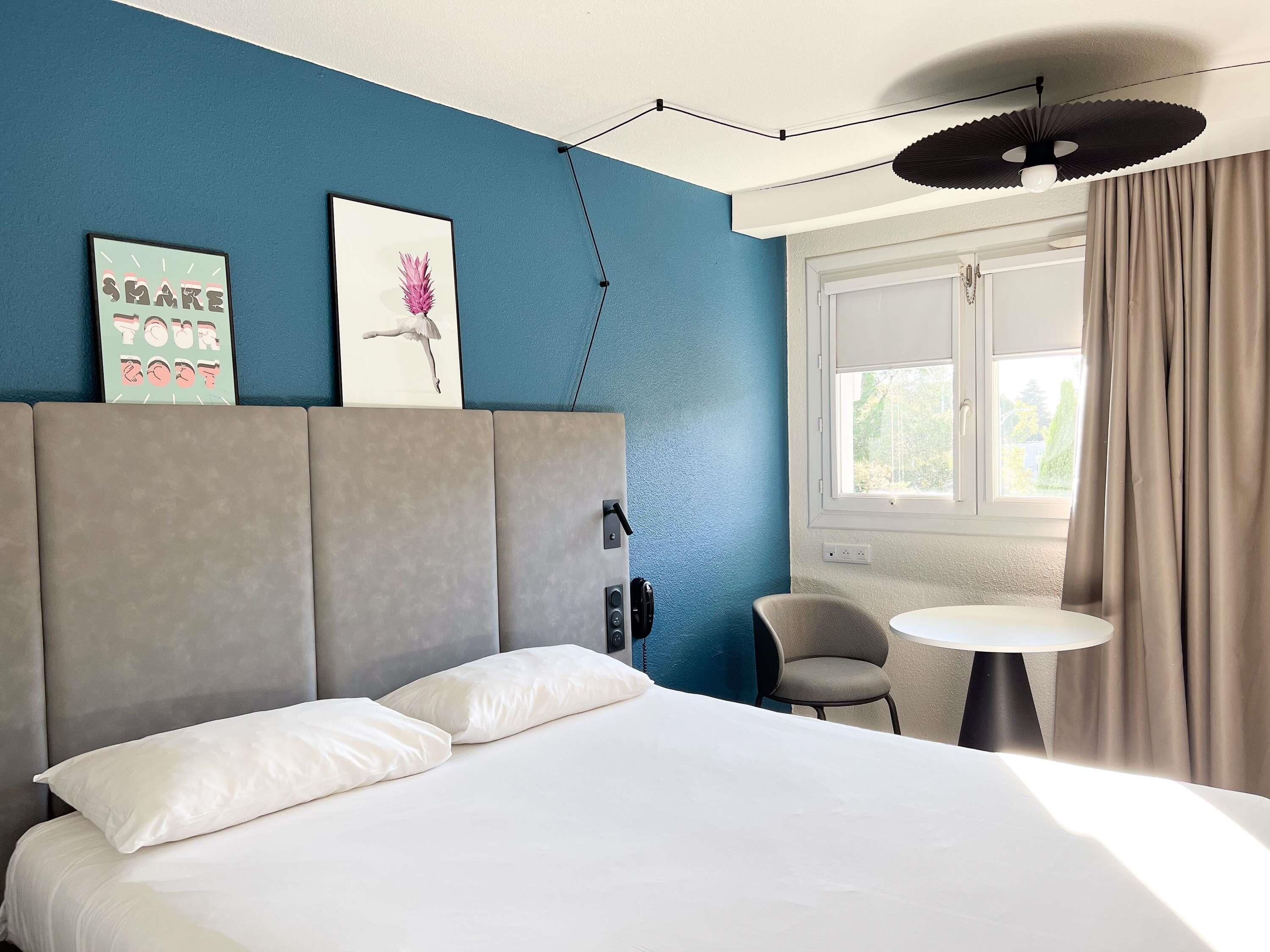 Ein Hotelzimmer mit einem großen Bett, einem kleinen Tisch, einem Stuhl, einem Kopfteil, einem Fenster mit Vorhängen und Wandbildern.