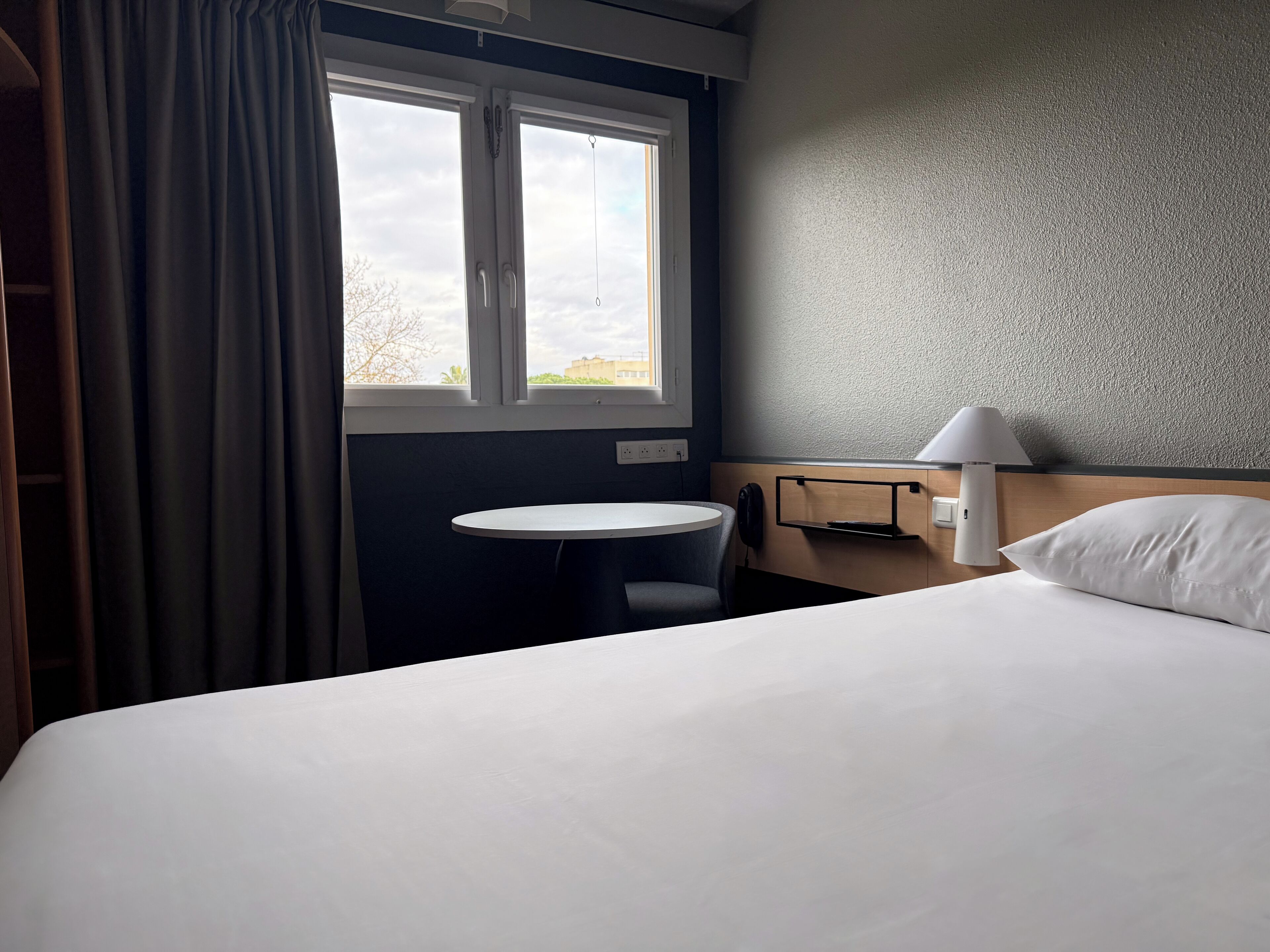 Habitació d'hotel amb llit, taula petita rodona, cadira, làmpada i finestra amb cortines.
