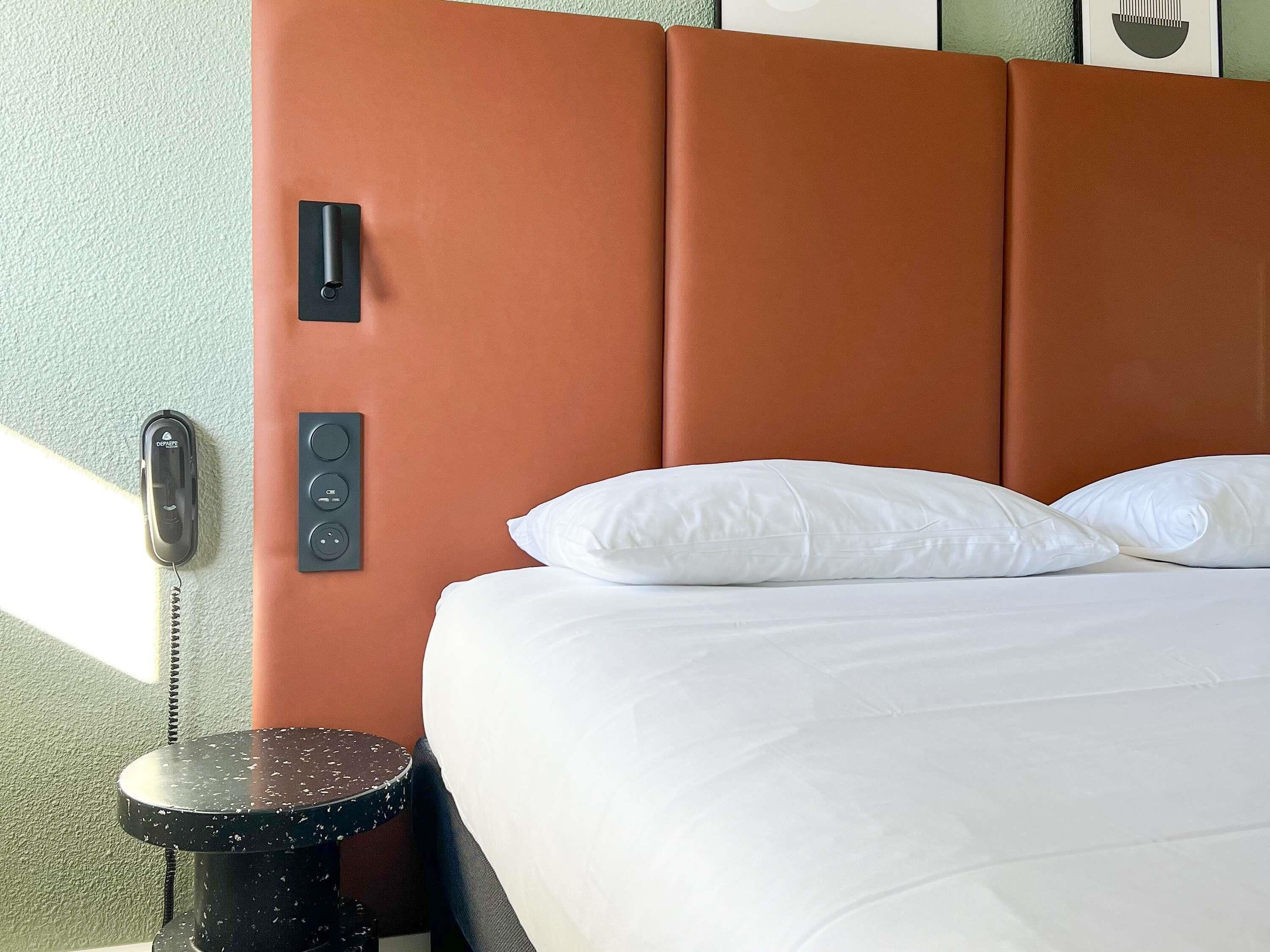 Una habitación de hotel con cama, almohadas blancas, un teléfono y una mesita.