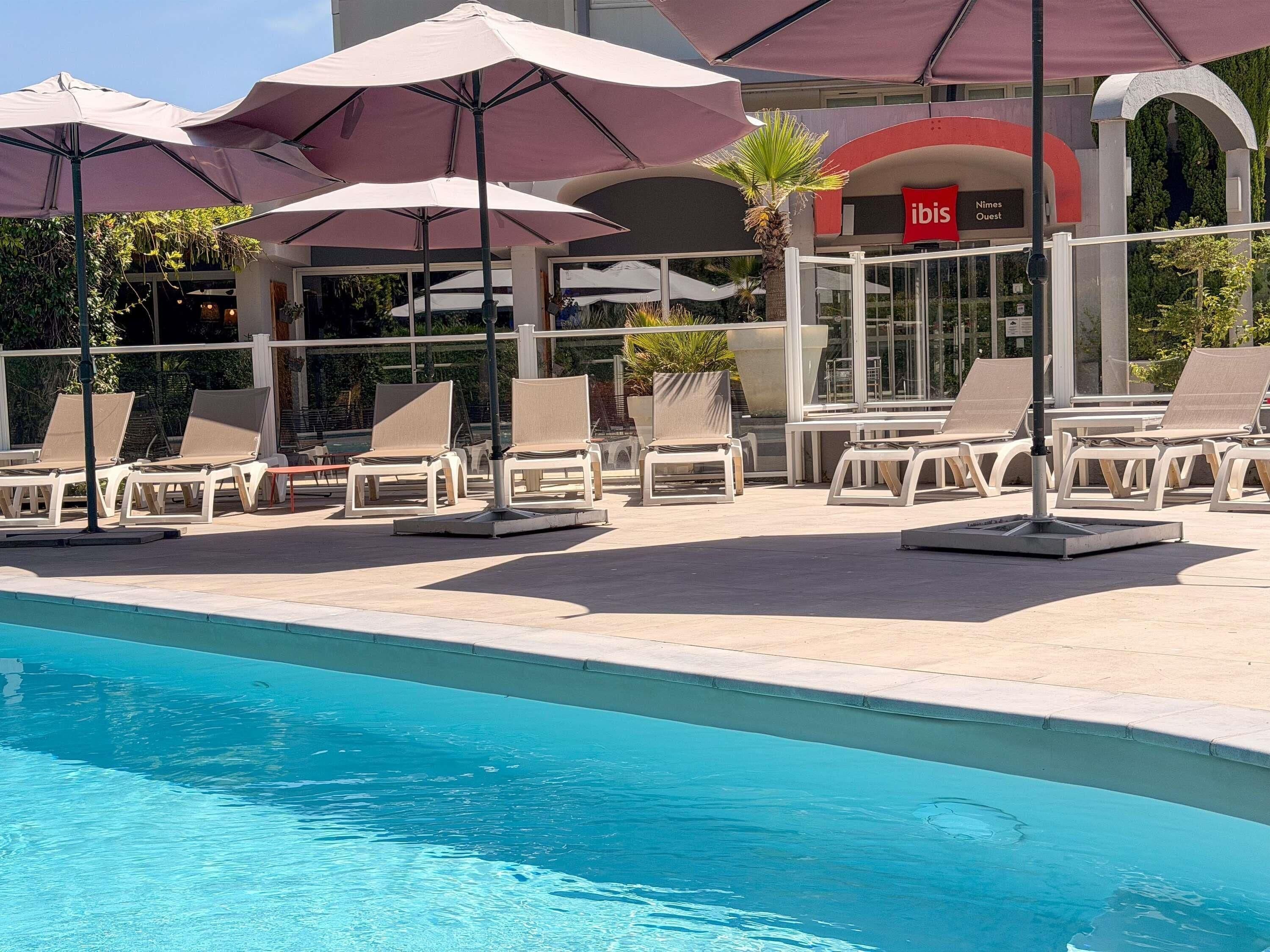 A poolside area with lounge chairs and umbrellas, a sign for 'ibis Nimes Ouest,' and a clear blue pool.