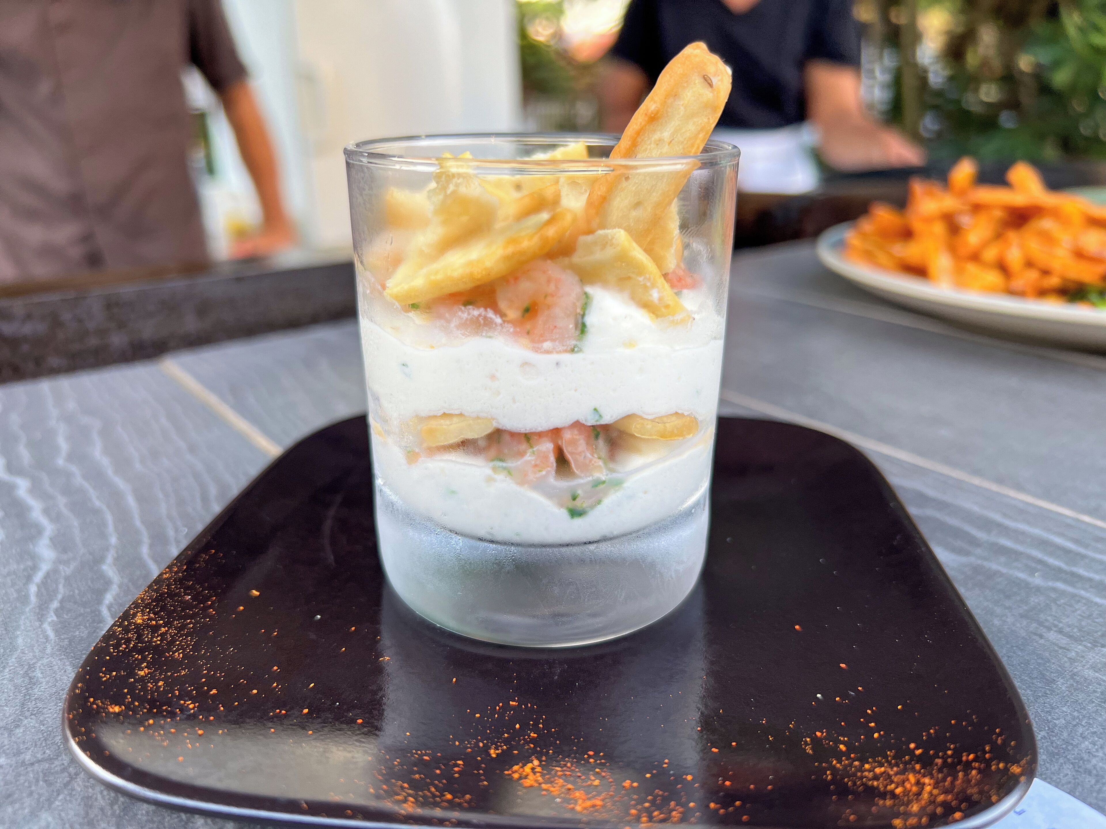 A dessert glass with layers of cream and fruit on a black square plate.