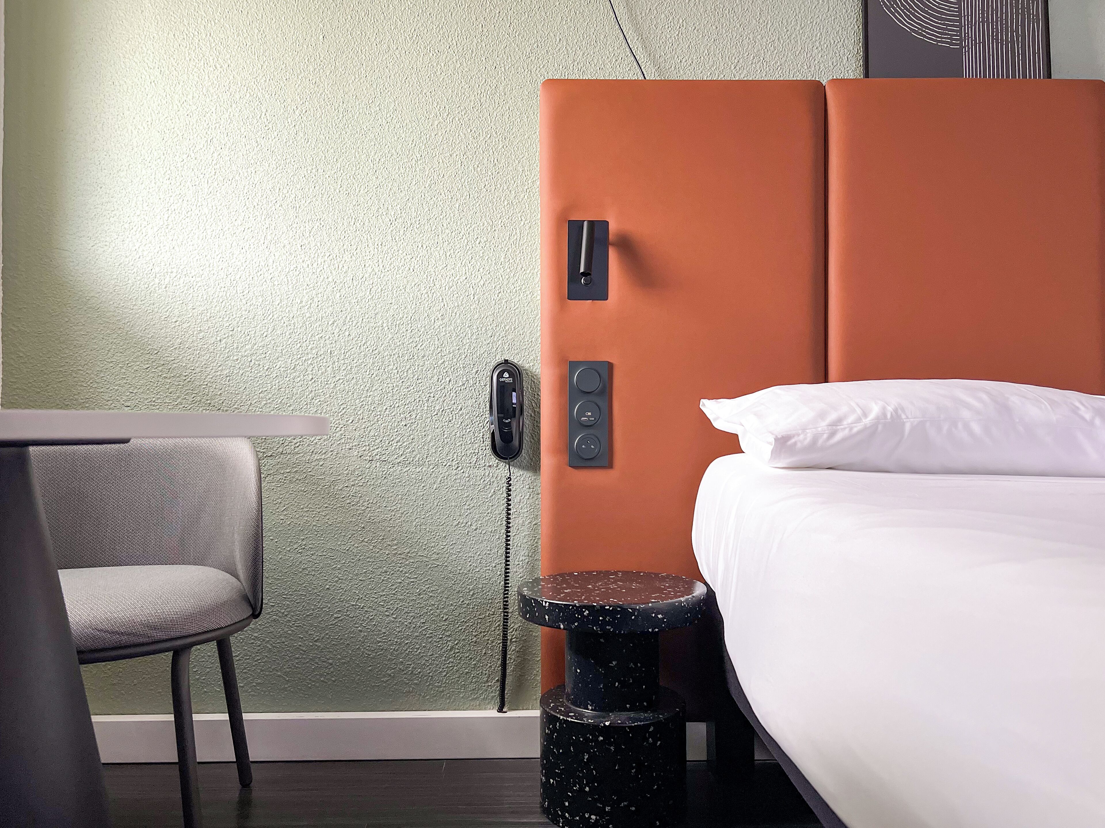 Habitación de hotel con cama, una silla, una mesita pequeña, teléfono y panel de control en la pared.