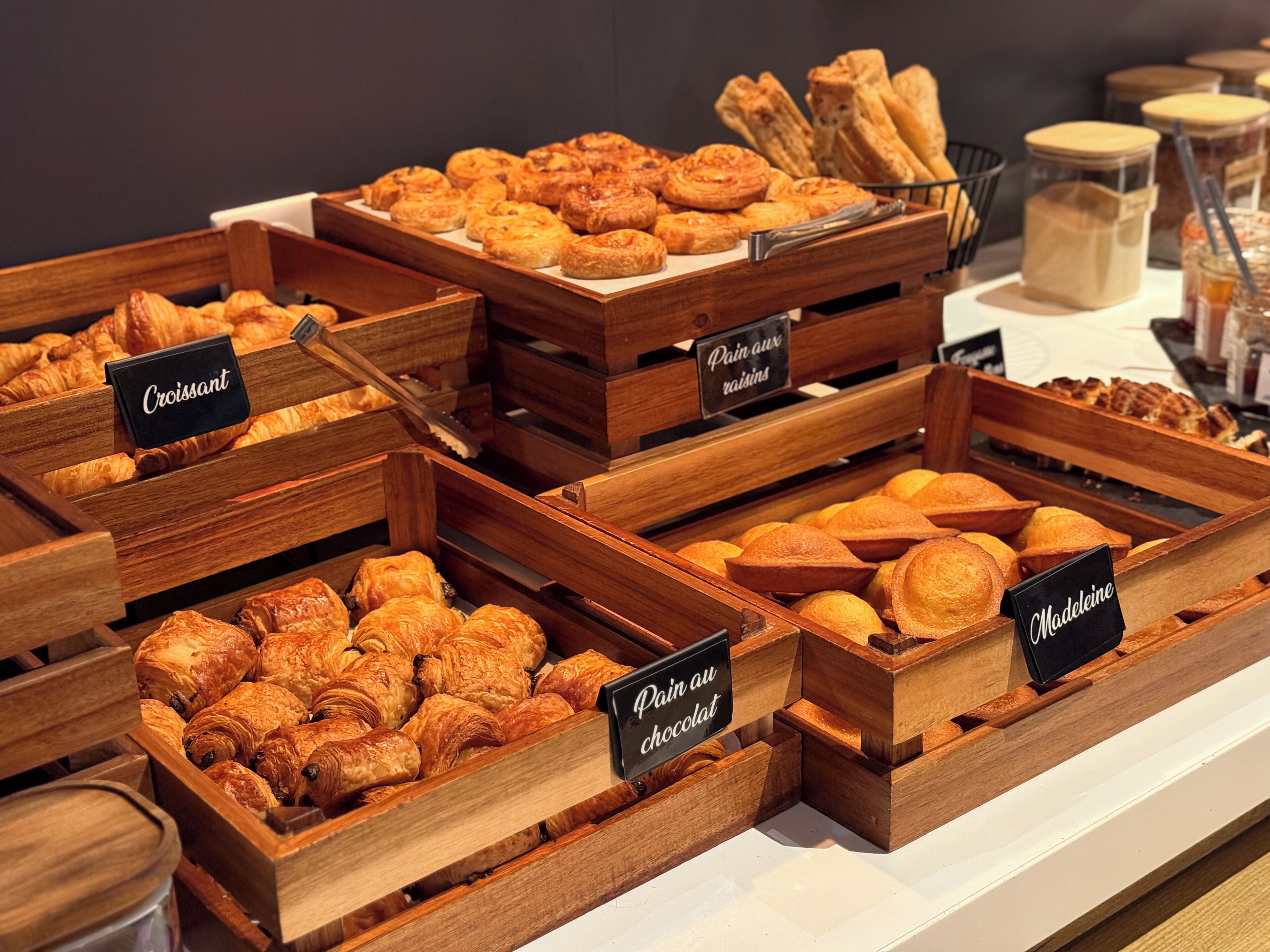 A bakery display with various types of pastries in wooden trays labeled with names like Croissant, Pain aux raisins, and Madeleine.