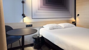 A hotel room with a bed, a desk, a chair, a wall-mounted picture, and a trash bin. - ibis Nimes Ouest (Nîmes)