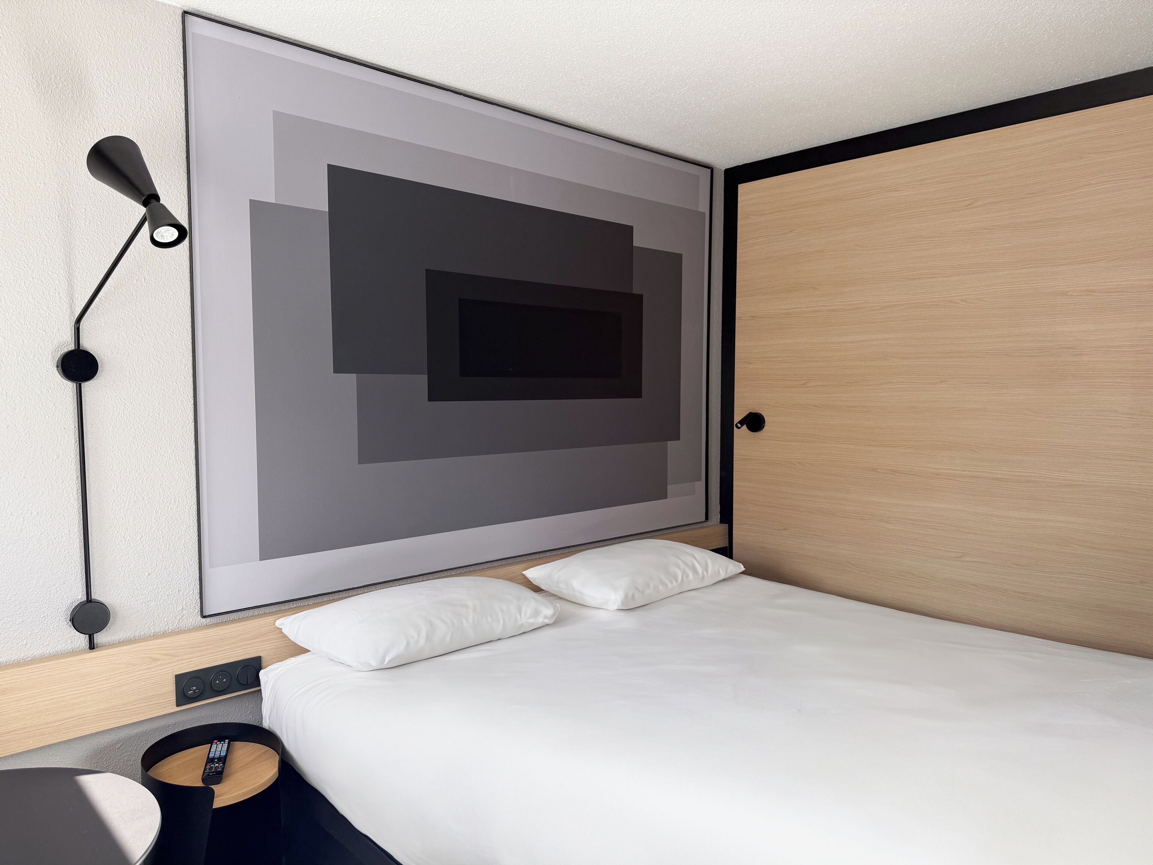 A modern hotel room with a large bed, a bedside table with a remote control, a wall-mounted lamp, and a large abstract artwork.