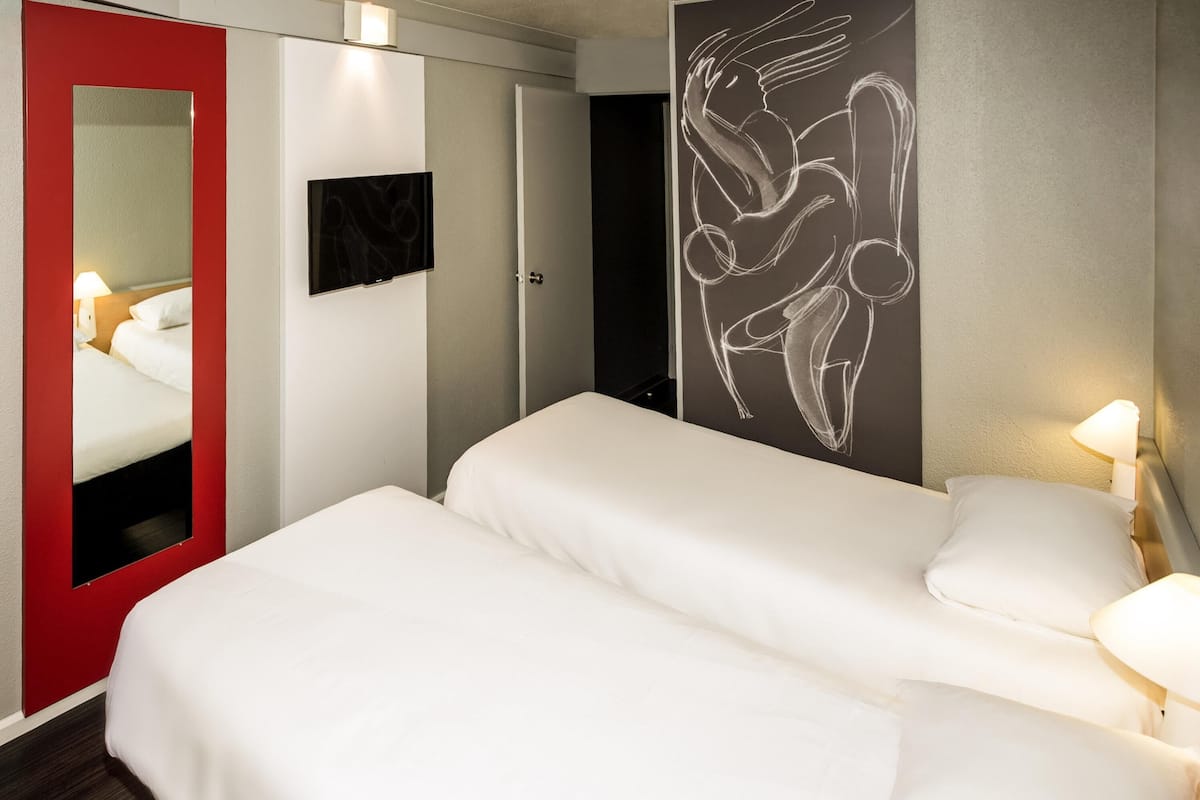 A hotel room with a bed, a red-framed mirror, a wall-mounted TV, and a wall art piece.