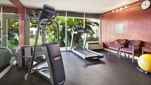 Ein Fitnessraum mit Laufbändern, einem Hometrainer und einem Rudergerät. Zwei Holzstühle und ein gelber Trainingsball sind ebenfalls vorhanden.