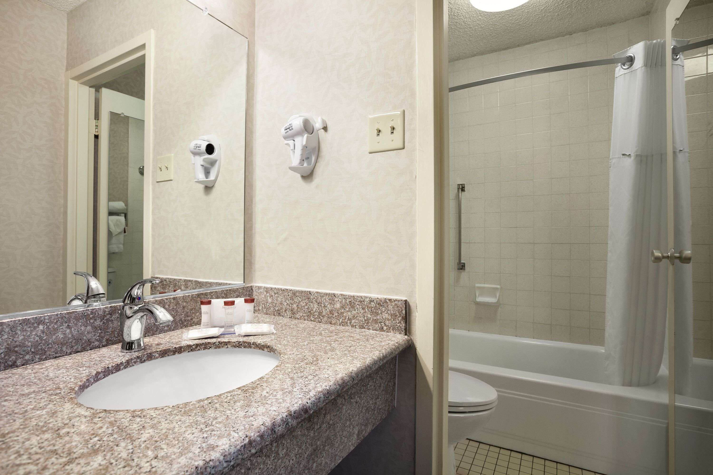 A bathroom with a granite countertop sink, a mirror, a hairdryer, and a shower area.