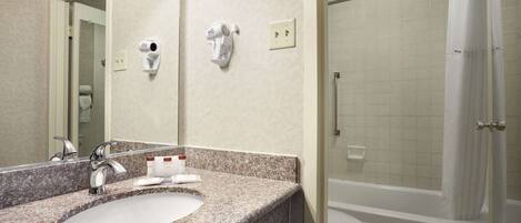 A bathroom with a granite countertop sink, a mirror, a hairdryer, and a shower area.