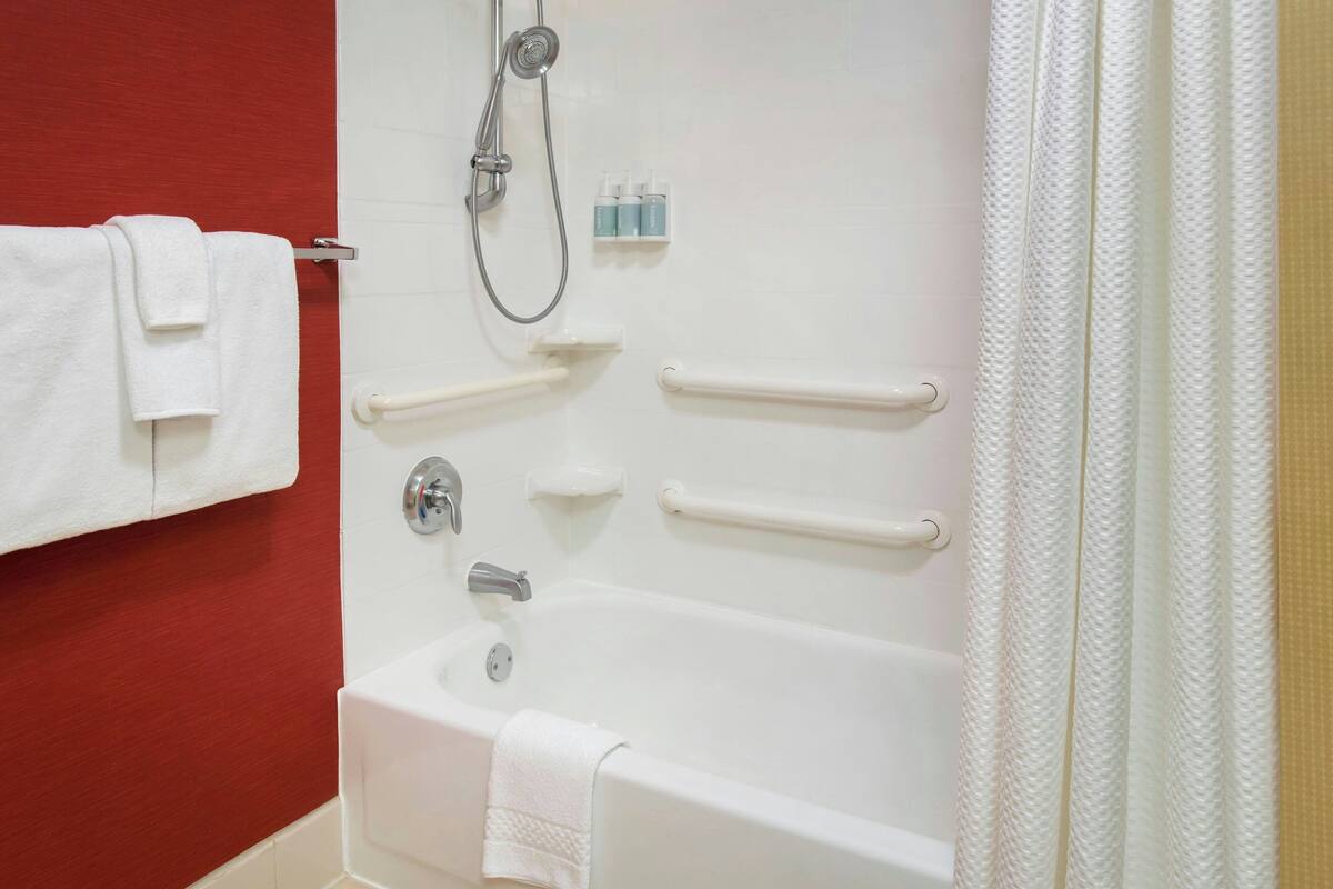 A bathroom with a white bathtub, a showerhead, handrails, and a towel rack.