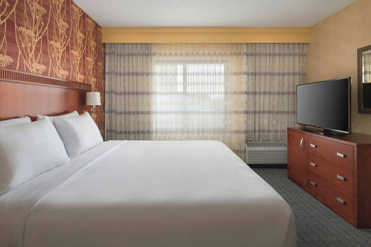 A hotel room with a large bed, a flat-screen TV, a wooden dresser, and a window with curtains.