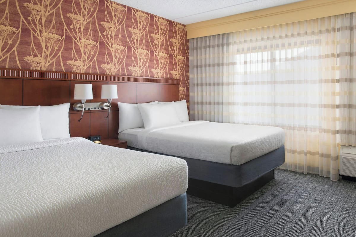 A hotel room with two beds, a headboard with a floral pattern, two bedside lamps, and a window with sheer curtains.