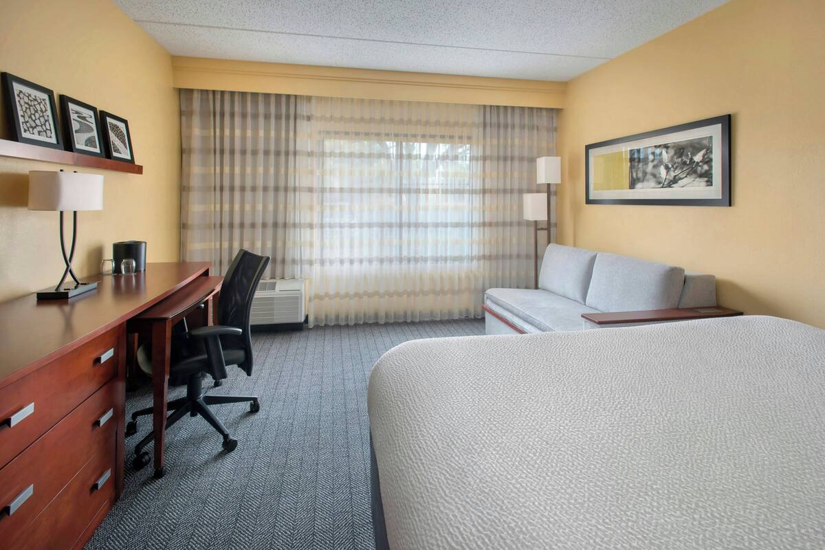 A hotel room with a bed, a desk with a chair, a lamp, and a window with curtains.